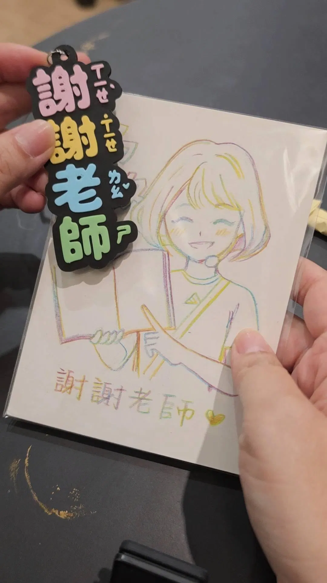 老師的彩色鉛筆似顏繪，畫中老師拿著書微笑，寫著「謝謝老師」，前面掛著 3D 列印的「謝謝老師」注音吊牌